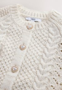 Cardigan en maille crème avec motifs torsadés et boutons en forme de cœur en perles, avec une étiquette de la marque NEXT à l'intérieur du col.