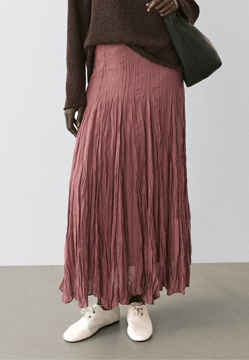 Personne portant une longue jupe froissée rose poussiéreuse, un pull marron, des chaussures blanches à lacets et tenant un sac à main noir sur un fond uni.