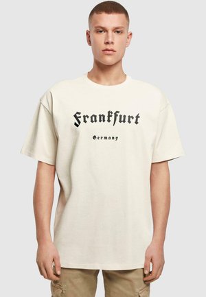 Beige oversized t-shirt made of cotton, featuring black text reading "Frankfurt" and "Germany" in a gothic font. Short sleeves, crew neck.