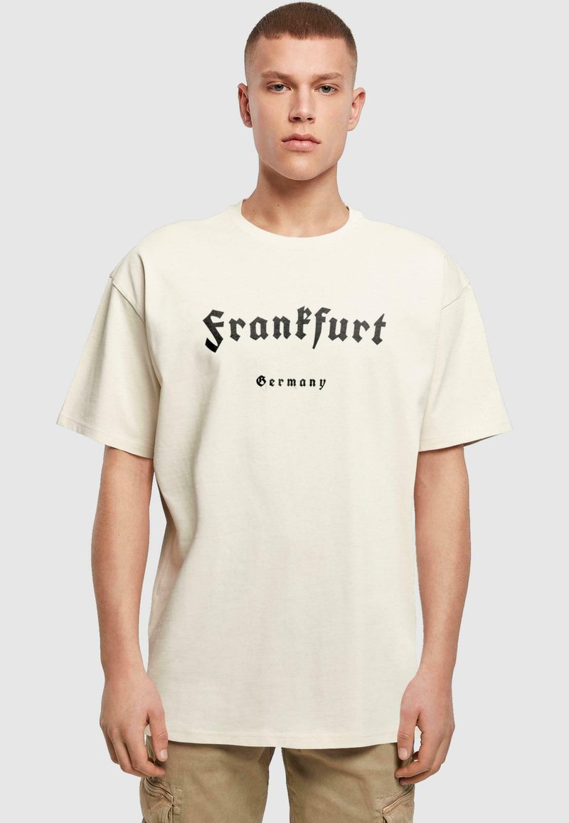 Beige oversized t-skjorte laget av bomull, med svart tekst som leser "Frankfurt" og "Tyskland" i en gotisk skrifttype. Korte ermer, rund hals.