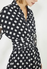 Zwarte jurk met een wit ruitpatroon, met kraagontwerp, vloeiende rok en textuurstof. Kenmerkt een getailleerde taille en knoopdetails.