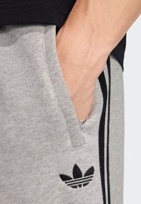 Sivé bavlnené tepláky s bočným vreckom, s čiernymi pruhmi a čiernym vyšitým logom na spodnom ľavom paneli.