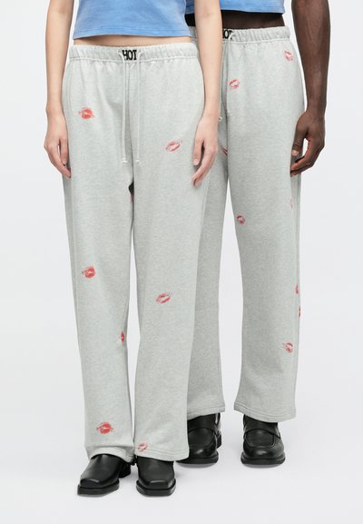 Divas personas valkā gaiši pelēkus sporta bikses ar sarkanu lūpu apdruku, saskaņotas ar melniem apaviem un īsiem ziliem topiem, stāv blakus viena otrai.