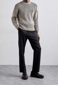 Beige gestreifter Strickpullover mit Rundhalsausschnitt, kombiniert mit schwarzen, maßgeschneiderten Hosen und schwarzen, klobigen Slippern, vor einem neutralen Hintergrund.