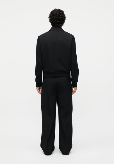 Άτομο με σγουρά σκουρόχρωμα μαλλιά στέκεται με την πλάτη, φοράει μαύρο μπουφάν και φαρδιά μαύρη παντελόνα μπροστά σε απλό φόντο.