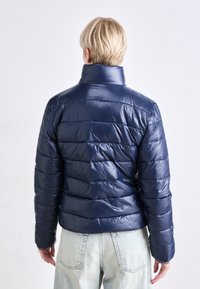 Námornícky modrá puffa bunda s prešívanou textúrou a vysokým golierom. Tkanina má lesklý povrch a rukávy sú zviazané elastickými manžetami.