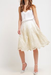 Witte camisole gecombineerd met een crèmekleurige rok met ingewikkelde gouden borduursels en een geschulpte zoom; glanzende zilveren hoge hakken.