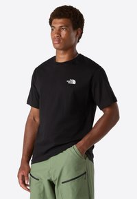 Man wearing black The North Face t-shirt and light green pants, standing with one hand in pocket, looking to the side against plain background.