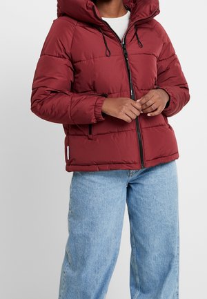 Persoon draagt een bordeauxrode gewatteerde jas met capuchon, zwarte ritssluiting en lichtblauwe wijde jeans, staand tegen een effen achtergrond.