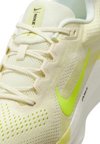 Primo piano di una scarpa da corsa Nike di colore crema, con tomaia in mesh, logo swoosh verde neon, lacci e scritta "AIR" sulla suola bianca.