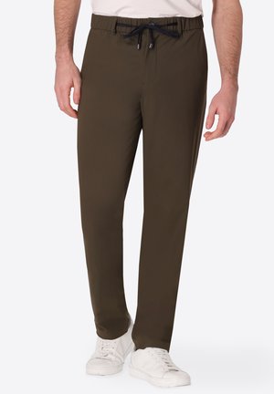Uomo che indossa pantaloni verde oliva con coulisse e sneaker bianche, in piedi davanti a uno sfondo chiaro e uniforme.