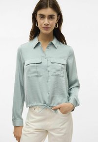 Camicia azzurra chiara con bottoni, due tasche sul petto e orlo con cordoncino, realizzata in un tessuto liscio e testurizzato. Maniche lunghe con polsini abbottonati.