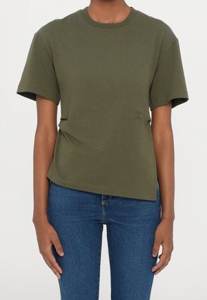 Person iført en ensfarvet olivengrøn kortærmet t-shirt og blå cowboybukser, stående med armene afslappet ned langs siderne.