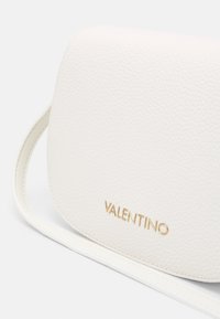 Bílá texturovaná kožená taška přes rameno s kulatým klopou, zdobená zlatým logem "Valentino" a tenkým bílým popruhem.