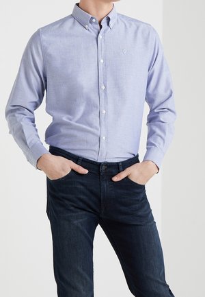 Homme portant une chemise à manches longues bleu clair et un jean bleu foncé, les mains dans les poches, debout contre un fond blanc.