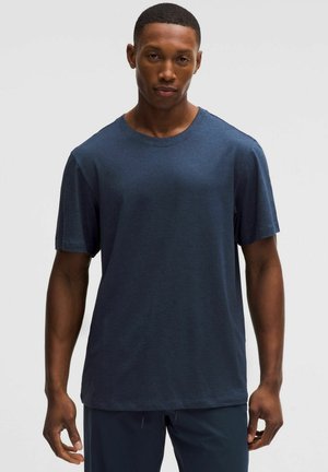 Uomo che indossa una t-shirt blu scuro a maniche corte tinta unita e pantaloni scuri, in piedi con le braccia rilassate davanti a uno sfondo chiaro e uniforme.