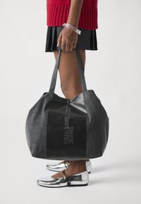 Grå tekstil tote bag med svart lærpanel som har embossed logo. Brede stropper og rommelig kropp. Sølvfargede sko synlig nedenfor.