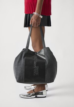 Grå tekstil tote bag med svart lærpanel som har embossed logo. Brede stropper og rommelig kropp. Sølvfargede sko synlig nedenfor.