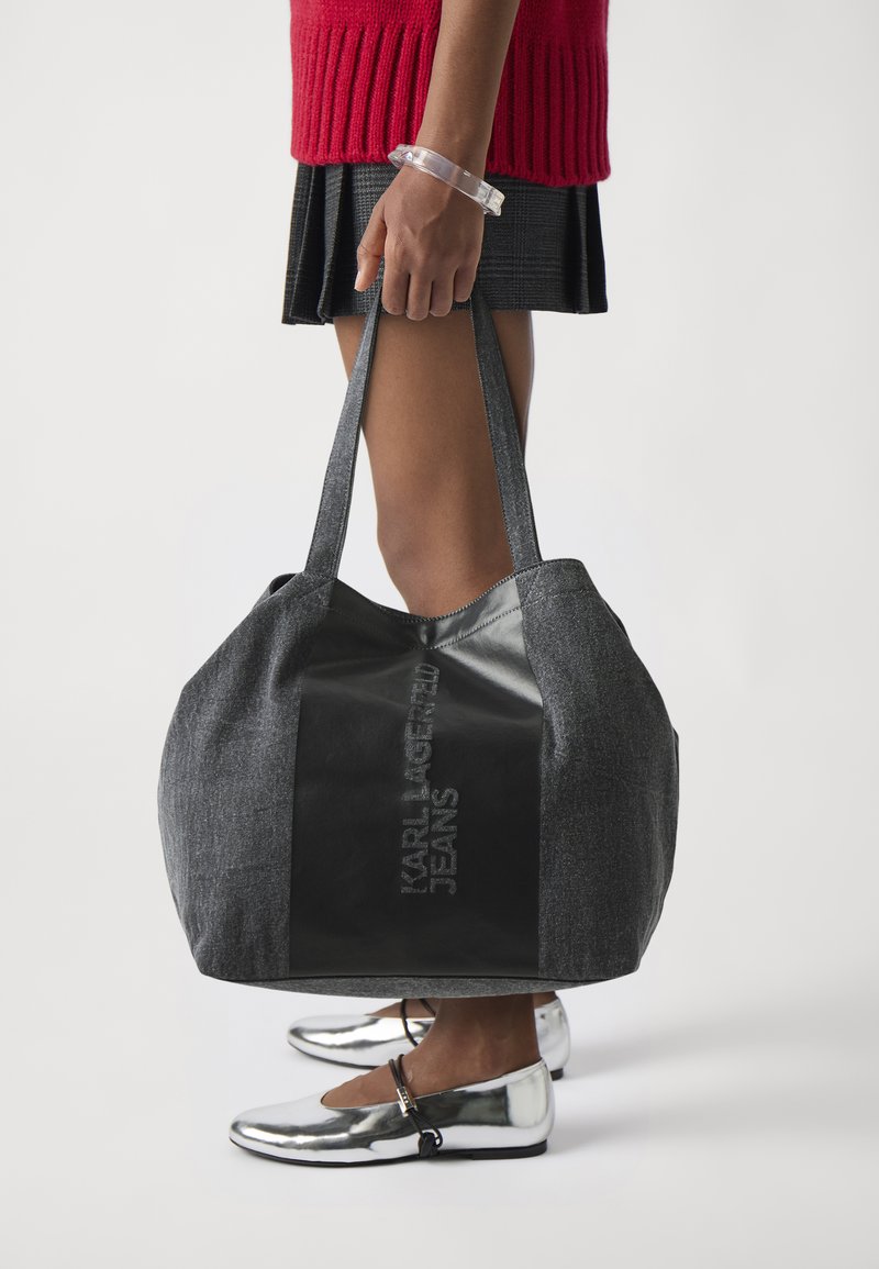 Grå tekstil tote bag med svart lærpanel som har embossed logo. Brede stropper og rommelig kropp. Sølvfargede sko synlig nedenfor.
