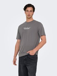 Junger Mann trägt ein graues T-Shirt mit dem Aufdruck "Sakura Blossom" und schwarze Jeans, steht mit einer Hand in der Tasche vor weißem Hintergrund.