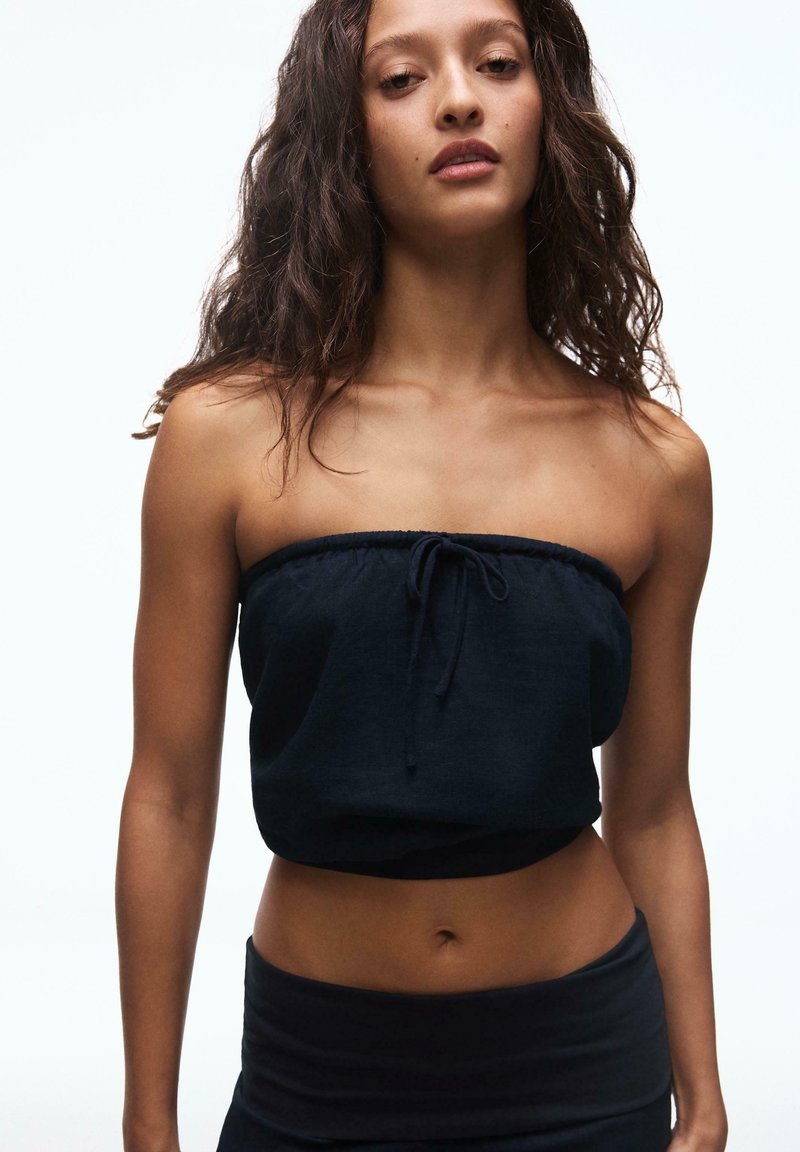 Jeune femme aux longs cheveux bouclés portant un haut court noir sans bretelles et un bas assorti, debout devant un fond clair uni.