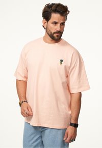 Rosa Baumwoll-T-Shirt mit lockerer Passform, das ein kleines gesticktes Ananasdesign auf der linken Brustseite zeigt. Kurze Ärmel und Rundhalsausschnitt.