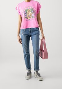 Persona vestida con una camiseta gráfica rosa con un leopardo y jeans azules, sosteniendo un bolso rosa, con zapatillas grises y una pulsera.