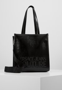 Błyszcząca czarna torba na ramię z podwójnymi uchwytami i regulowanym paskiem, z napisem "Versace Jeans Couture" wytłoczonym z przodu.