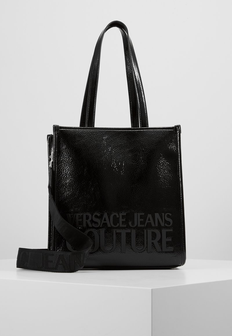 Błyszcząca czarna torba na ramię z podwójnymi uchwytami i regulowanym paskiem, z napisem "Versace Jeans Couture" wytłoczonym z przodu.