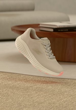 Sneaker sportiva beige con tomaia in mesh e suola texturizzata che galleggia sopra un pavimento moquettato in un soggiorno con libri su un tavolino rotondo di legno.