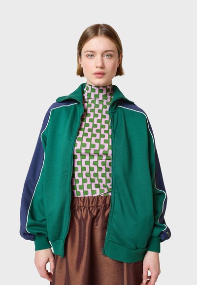 Grüne und marineblaue Zip-Jacke mit weißen Piping-Details. Getragen über einem rosa und grünen gemusterten Rollkragenpullover. Weicher Stoff und lockere Passform.