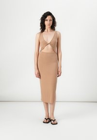 Robe en tricot beige côtelé avec un décolleté en V profond et un design découpé à la taille, s'étendant jusqu'à la longueur midi. Associée à des tongs noires.