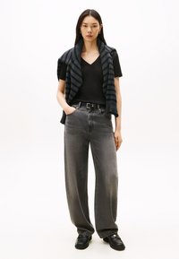 Nő fekete V-nyakú pólóban, sötétszürke csíkos pulóvert vállán keresztben viselve, széles szürke farmert, fekete övet és fekete mokaszint visel, egyik kezét zsebében tartva áll.