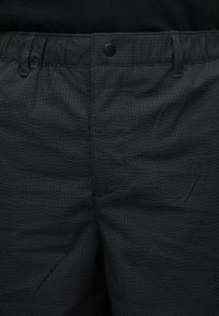 Nærbilde av teksturerte svarte bukser med knepp-knapp lukking og beltestropper, brukt med en svart topp.