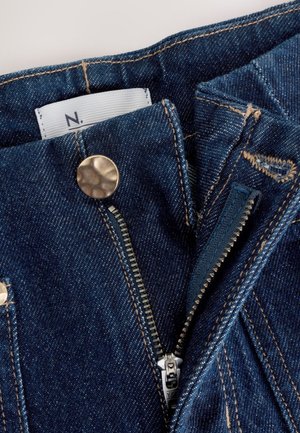 Blugi din denim de culoare albastru închis, cu fermoar metalic și etichetă vizibilă în interior. Cusăturile sunt într-o nuanță mai deschisă, creând un contrast.