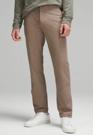 Beżowe dopasowane spodnie o gładkiej fakturze, o wąskim kroju i delikatnych załamaniach, zestawione z białymiSneakerami oraz oliwkowym topem z długim rękawem.