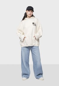 Sudadera holgada color crema con un bolsillo frontal y cuatro parches gráficos; combinada con jeans de pierna ancha color azul claro y zapatillas blancas. Lleva puesta una gorra negra.