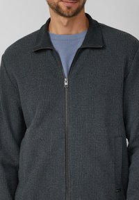 Graue Jacke mit Reißverschluss aus strukturiertem Stoff mit einem gerippten Design. Verfügt über einen hohen Kragen und einen silbernen Reißverschluss.
