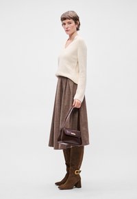 Μπέζ πλεκτό πουλόβερ, καφέ φούστα με πτυχώσεις, καφέ σουέτ μπότες μέχρι το γόνατο με χρυσές λεπτομέρειες και καφέ τσάντα χειρός με διακριτική μεταλλική λεπτομέρεια.