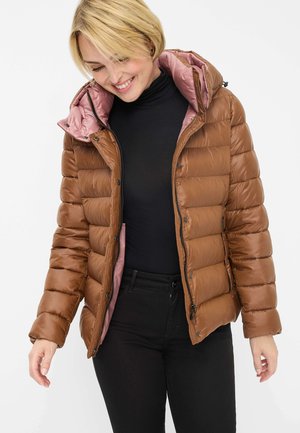 Frau trägt eine braune gesteppte Jacke mit pinker Innenfutter über einem schwarzen Rollkragenpullover und schwarzen Hosen, lächelt und schaut nach unten.
