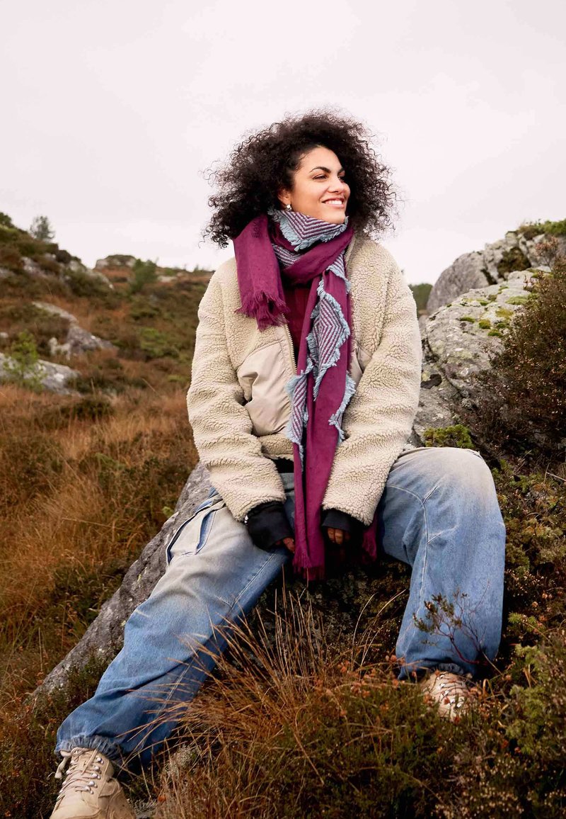 Giovane donna con capelli ricci seduta su una roccia all'aperto, indossa un giubbotto di pile beige, una sciarpa viola, jeans larghi e scarponi da trekking beige.