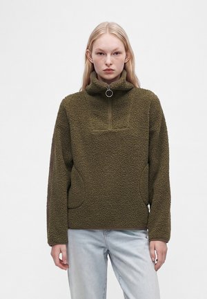 Femme portant un pull polaire vert olive avec fermeture éclair devant et un jean bleu clair sur un fond uni.
