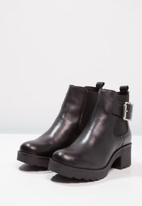 Botas de cuero negras hasta el tobillo con un tacón grueso, acabado texturizado, paneles laterales elásticos y un detalle de hebilla de metal en el lado exterior.