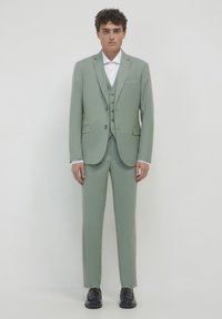 Completo tre pezzi di colore verde chiaro con un design slim fit, caratterizzato da rever a punta, gilet con bottoni e pantaloni affusolati. Scarpe nere completano l'outfit.