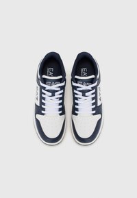 Hvite og marineblå sneakers med perforert tåområde, hvite snørebånd og detaljering av logo på siden. Glatt lær og stoffmaterialer. Flat såle.