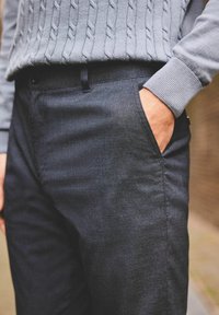 Donker gemusterde broek met een textuurafwerking. Voorzien van een zijzak en een relaxte pasvorm. Gecombineerd met een lichtgrijze kabelgebreide trui.