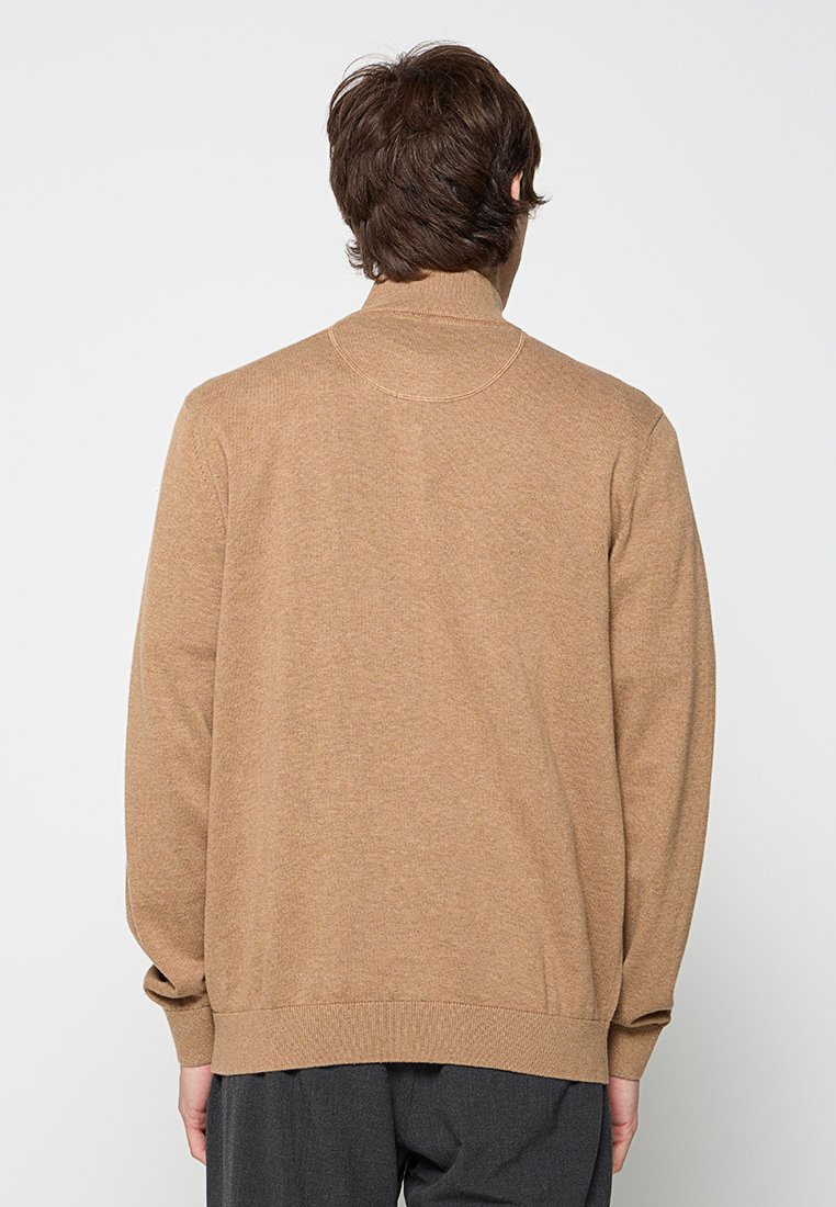 Mann in einem schlichten, camel-farbenen Pullover und dunklen Hosen, von hinten vor einem schlichten, hellen Hintergrund gezeigt.