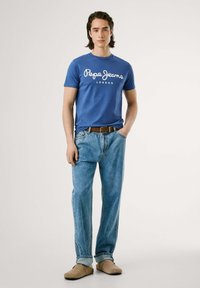 Tricou din bumbac bleu cu logo-ul alb "Pepe Jeans London", purtat cu blugi denim deschiși la culoare și pantofi slip-on bej. Design simplu și casual.