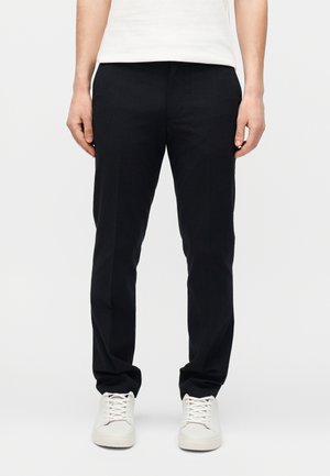 Mand iført sorte bukser med lige ben og hvide sneakers, stående mod en ensfarvet hvid baggrund.