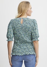 Bluse mit floralem Muster in Grün und Blau, mit Puffärmeln, geriffelter Textur und einem Schlüsseloder-Detail am Rücken. Kombiniert mit blauen Jeans.